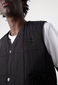 Vest negru, matlasat, cu nasturi tip snap și un finisaj texturat, având un logo ridicat de culoare neagră "HUGO" pe partea stângă a pieptului. Purtați peste o cămașă albă.