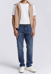Gilet a maglia beige chiaro sopra una t-shirt bianca, abbinato a jeans di denim blu e sneakers bianche. Look casual e design semplice.