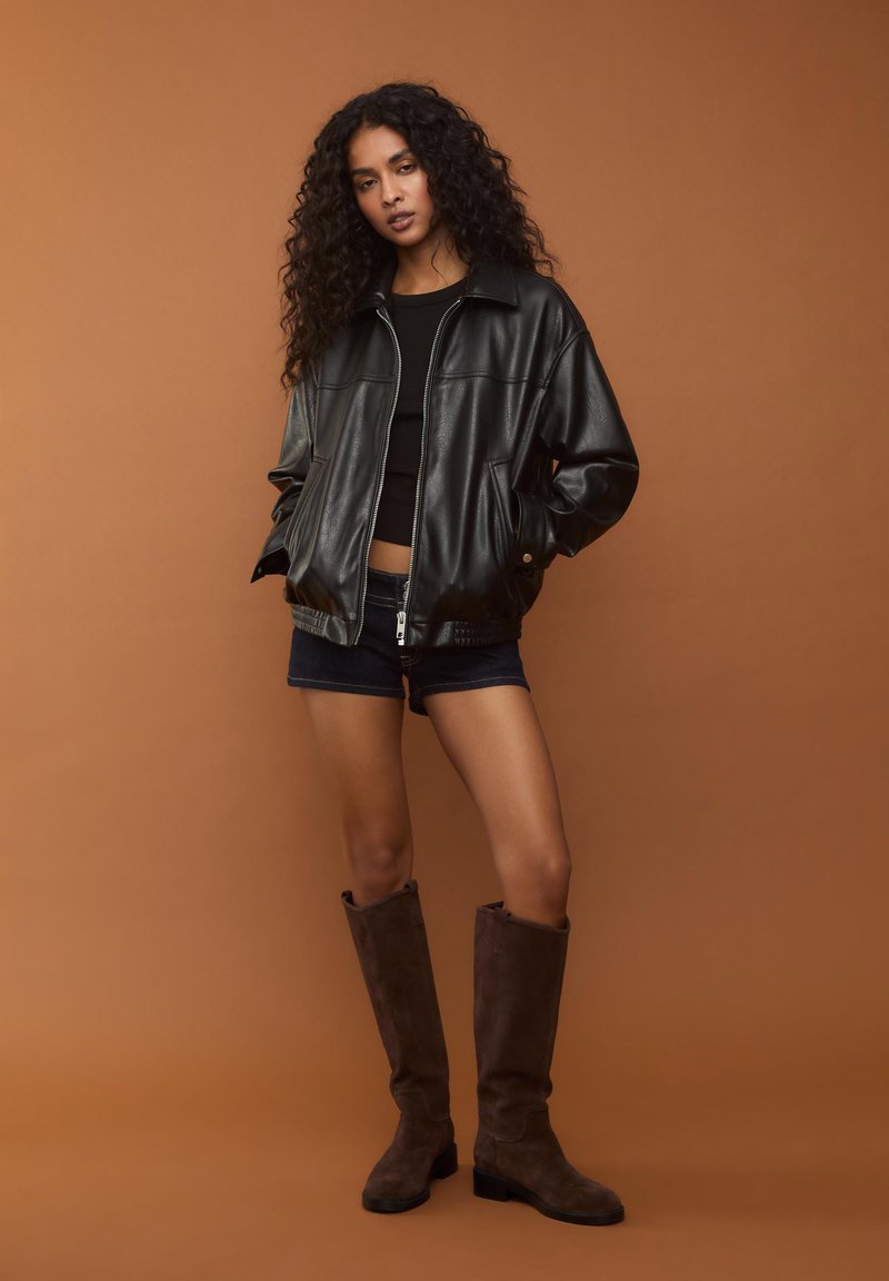 Giubbotto bomber in pelle nera con zip frontale, indossato sopra una maglietta nera attillata. Abbinato a pantaloni corti in denim scuro e stivali alti fino al ginocchio in suede marrone.
