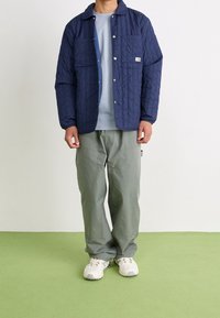 Marinblå quiltad jacka med tryckknappar och två bröstfickor, i kombination med en ljusblå skjorta och olivgröna byxor, fulländat med vita sneakers.