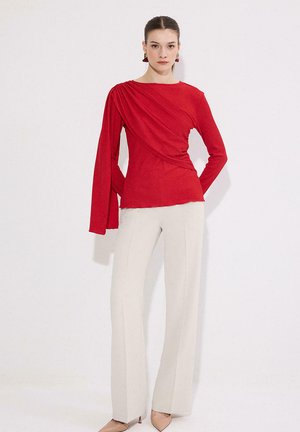 Žena v červenom asymetrickom top s dlhým rukávom a drapovaným detailom na jednej ruke, v kombinácii s krémovými širokými nohavicami s vysokým pásom a béžovými lodičkami.