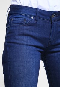 Jeans de denim azul oscuro con un diseño clásico de cinco bolsillos, herrajes metálicos y un ajuste ajustado con una textura suave.