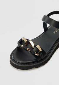Schwarze Sandale mit dickem Sohle, doppeltem Riemendesign und Kettendetail mit goldenen Akzenten. Glatte Lederoberfläche, minimalistischer Look.