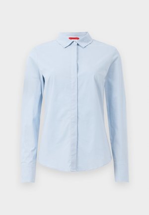 Chemise bleu clair à manches longues en coton, avec un col à boutons, une patte de boutonnage à l'avant et des manches à boutons. Elle présente une texture lisse.