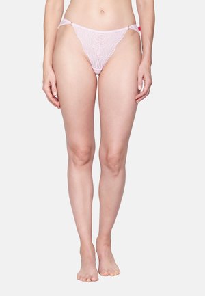 Woman wearing light pink sheer lace bikini-style underwear, standing barefoot against a plain light background.