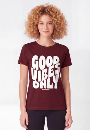 Junge Frau mit lockigem Haar, die ein kastanienbraunes Shirt mit großem weißen Text "GOOD VIBES ONLY" auf schlichtem Hintergrund trägt.