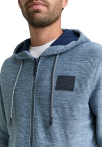 Brodaty mężczyzna ubrany w teksturowaną niebieską bluzę na zamek z białymi i niebieskimi sznurkami oraz ciemną łatą na klatce piersiowej, na białym t-shircie.