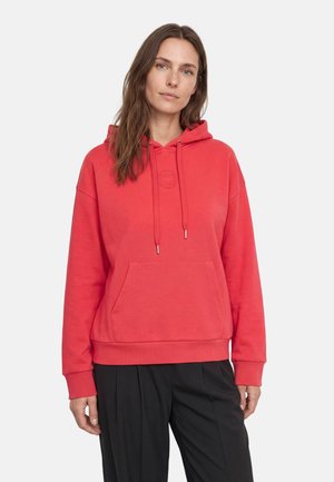Donna che indossa una felpa rossa con cappuccio e tasca frontale e pantaloni neri, in piedi davanti a uno sfondo chiaro e uniforme.