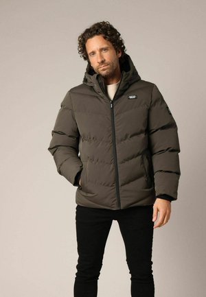 Doudoune vert olive avec capuche. Comprend une fermeture éclair à l'avant, un design matelassé et des accents noirs. Portée avec un pantalon noir.