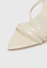 Sandale à talon haut en cuir verni beige avec un bout pointu et trois brides plates traversant le pied. Texture lisse et design élégant.