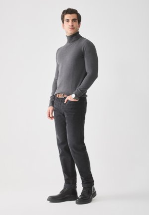 Grijze coltrui, nauwsluitend ontwerp, gedragen met zwarte jeans en zwarte laarzen. Model staat met handen in de zakken tegen een lichte achtergrond.