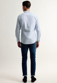 Camisa de rayas azul claro de manga larga con puños abotonados, combinada con jeans ajustados azul oscuro y zapatos negros, vista desde atrás.