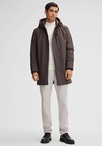 Mann in einem dunkelbraunen winterlichen Kapuzenmantel, hellgrauen Hosen, einem weißen Pullover und schwarzen Lederschuhen, der vor einem schlichten Hintergrund steht.