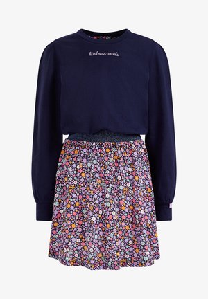 Barn iført navyblå langærmet skjorte med teksten "kindness counts", blomsterprintet nederdel, lilla sokker og skinnende lyserøde snørestøvler, stående.