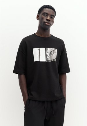 Giovane uomo con capelli corti e ricci indossa una maglietta nera oversize con una foto urbana in bianco e nero e la parola "LOCK" sul davanti. Mani in tasca.
