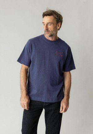 Mand med kort brunt hår og skæg iført en navyblå t-shirt og sorte jeans mod en ensartet lys grå baggrund.