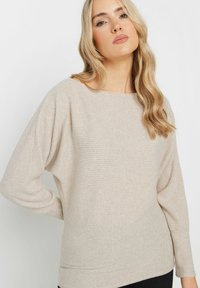 Beige ribbknitted neulepaita, jossa on leveä kaula-aukko ja roikkuva olkamuoto. Pehmeä rakenne ja hienovaraiset pystysuorat raidat. Rennon istuvuuden.