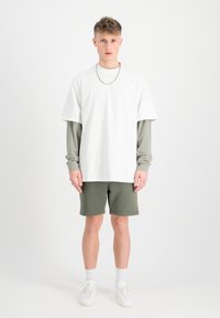 T-shirt blanc à manches courtes sur une chemise à manches longues verte, shorts olive et baskets blanches. Collier en chaîne argentée ; look décontracté en superposition.