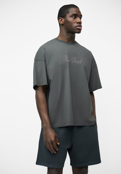 Jeune homme portant un t-shirt ample gris foncé avec un texte discret « Pale Mind » et un short foncé, debout devant un fond blanc uni.