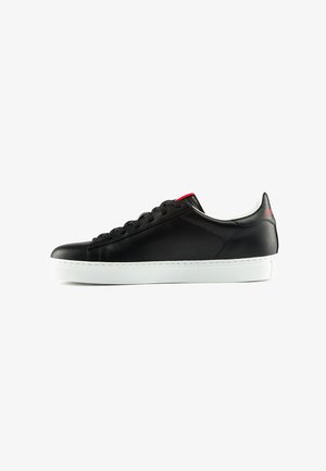 Zapatillas de cuero negro con un acabado suave, suela de goma blanca, puntera redondeada y cordones negros; presenta un acento rojo cerca de los ojales.