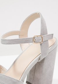 Sandale à talon haut en daim gris clair avec un talon épais et une bride ajustable à la cheville avec une petite boucle dorée.