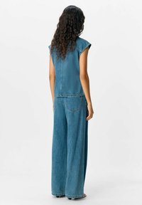 Monos de mezclilla en azul medio, con mangas cortas tipo capa, pantalones de pierna ancha, bolsillos traseros y costuras visibles a lo largo de las costuras.