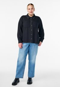 Čierna košeľa s gombíkmi a dvoma vreckami na hrudi, s bielymi švami, kombinovaná so svetlomodrými širokými džínami a čiernymi členkovými čižmami.