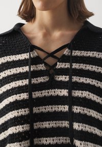 Femme portant un haut en maille rayé noir et blanc avec un col à lacets croisés et un col, dévoilant son cou et ses épaules.