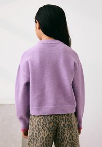 Lila gestrickter Pullover mit rundem Halsausschnitt und geripptem Saum, kombiniert mit beige-farbenen, hochtaillierten Hosen mit Leopardenmuster, von hinten gezeigt.