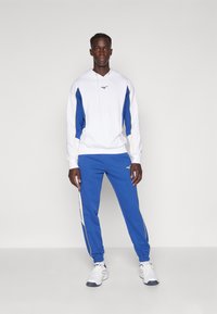 Hvit hettegenser med blå sidestykker og gule detaljer, sammen med blå joggebukser med hvite striper og elastiske mansjetter, brukt med hvite sneakers.