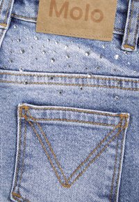 Un jean en denim bleu clair présente une poche avec des coutures dorées et des embellissements de petits rhinestones regroupés en motifs. Étiquette de la marque Molo visible.