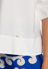 Blouse en coton blanc avec une étiquette visible à l'ourlet. La blouse possède des manches courtes et une texture lisse et légère.