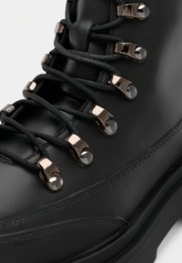 Fekete bőr túrabakancs fémbetétellel és vastag fekete fűzőkkel. Fényes díszítések és texturált varrás a szélek mentén.
