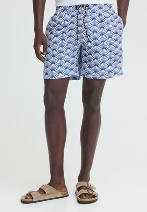 Man draagt blauw-witte korte broek met patroon, handen in zakken, gecombineerd met beige sandalen met dubbele bandjes tegen een effen achtergrond.