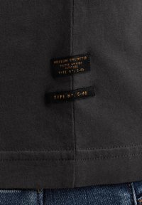 Dark grey cotton t-shirt fabric with ribbed texture. Two black label tags with gold text, stitched on the lower hem.