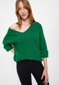 Grüner gestrickter Pullover mit tiefem V-Ausschnitt, hochgerollten Ärmeln und strukturiertem Muster; kombiniert mit schwarzen, figurbetonten Jeans.