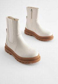 Bottes chelsea en cuir avec une tige beige, panneaux latéraux élastiques, fermeture à glissière et une semelle en caoutchouc marron au design texturé et robuste.