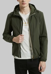 Veste légère verte avec capuche, poignets élastiques et fermeture éclair sur le devant. Portée sur un t-shirt blanc avec texte jaune. Texture simple et lisse.