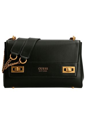 Sac à main Guess en cuir noir avec bandoulière chaîne dorée, détails de boutons-pression ton or, et logo "GUESS EST 1981" sur le rabat avant.