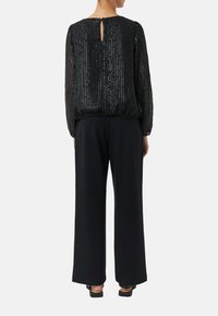 Schwarze, strukturierte Bluse mit vertikalen Streifen, Detail mit Schlüsselloch am Rücken; kombiniert mit locker sitzenden schwarzen Hosen. Akzentuiert mit glänzendem Stoff.