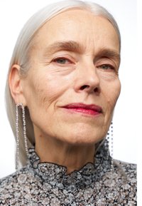 Gros plan sur une personne avec des cheveux argentés, portant un blouse noire à motifs floraux avec un col volé et de longues boucles d'oreilles argentées qui pendent. Peau lisse.