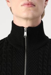 Pull en maille noire avec un col haut côtelé. Présente une fermeture éclair argentée et des motifs texturés, y compris des torsades et des points.
