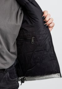 Chaqueta de plumas negra con un bolsillo con cremallera en el interior a la izquierda. La parte exterior es lisa, mientras que el forro es de un gris claro contrastante.