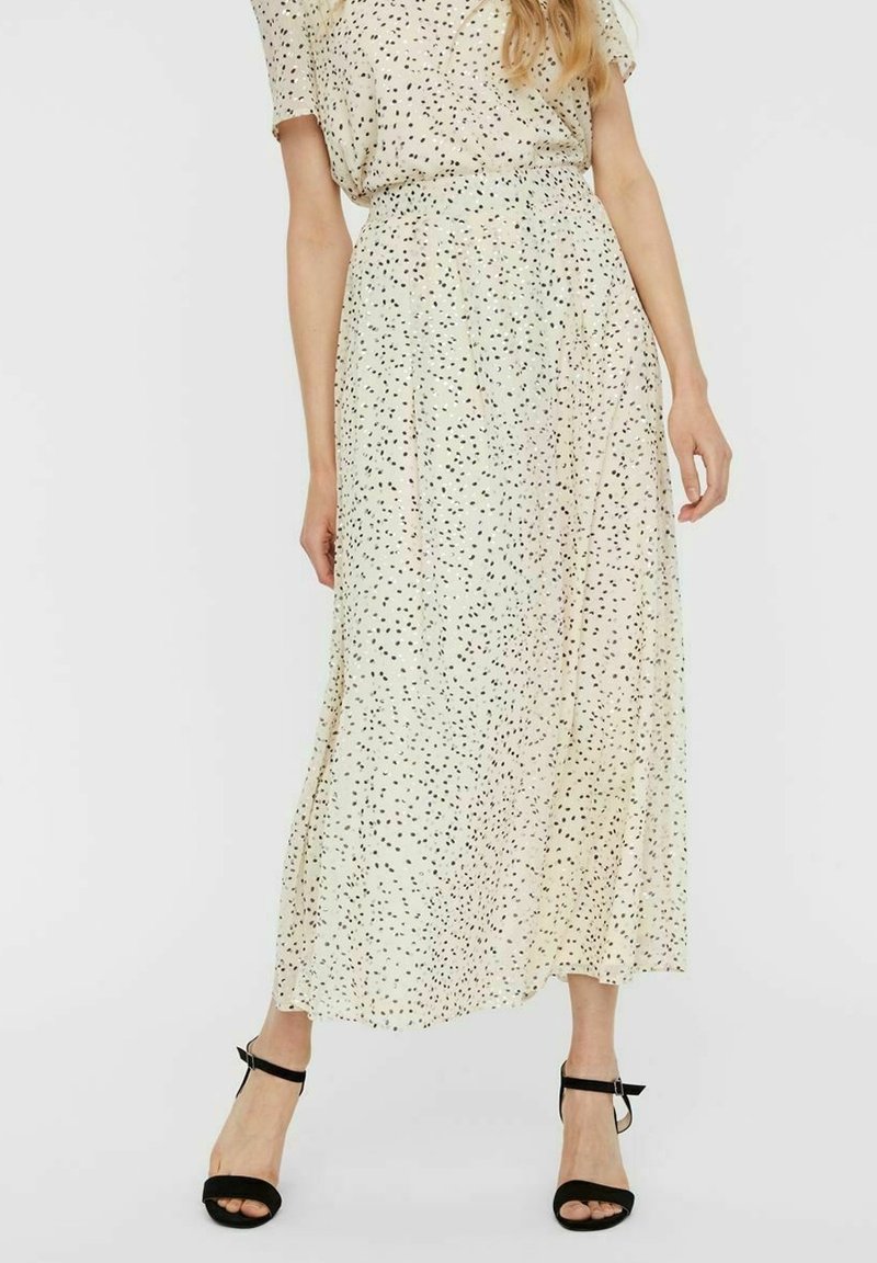 Jupe longue en tissu beige clair avec des éclaboussures noires éparses. Dispose d'une taille élastiquée et tombe jusqu'aux chevilles. Portée avec des sandales noires à brides.