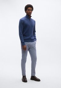 Μπλε πουλόβερ με πλεκτό σχέδιο cable και ψηλό γιακά, συνδυασμένο με ανοιχτό γκρι παντελόνι και σκούρα καφέ παπούτσια. Το σχέδιο είναι υφασμένο και εφαρμοστό.