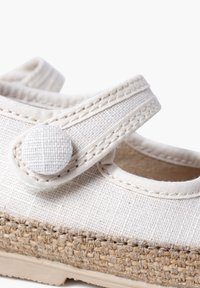 White canvas shoe with a textured strap and round button closure; features a braided jute midsole and a smooth rubber outsole.
