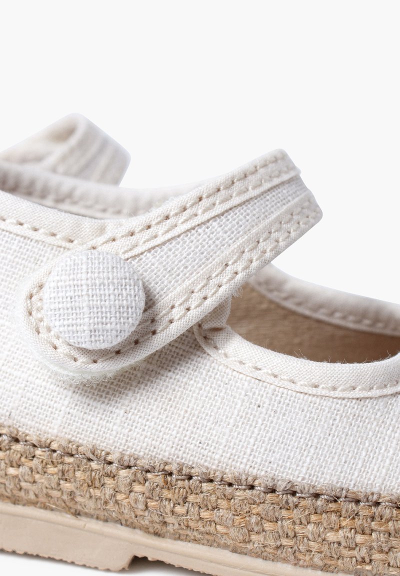 White canvas shoe with a textured strap and round button closure; features a braided jute midsole and a smooth rubber outsole.