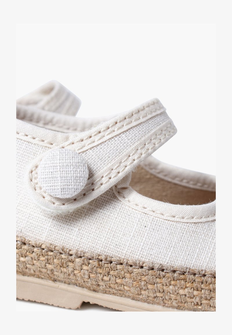 Zapato de lona blanco con una correa texturizada y cierre de botón redondo; cuenta con una entresuela de yute trenzado y una suela de goma suave.