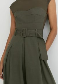 Olivekroheline kleit, millel on vormitud bodi, kapuutsiga varrukad ja lai vöö. Plisseeritud seelik lisab mahtu, kangas on ühtlase tekstuuriga.