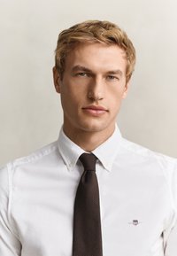 Jeune homme aux cheveux blonds courts et aux yeux bleus, portant une chemise blanche boutonnée et une cravate sombre, regardant droit devant avec une expression neutre.